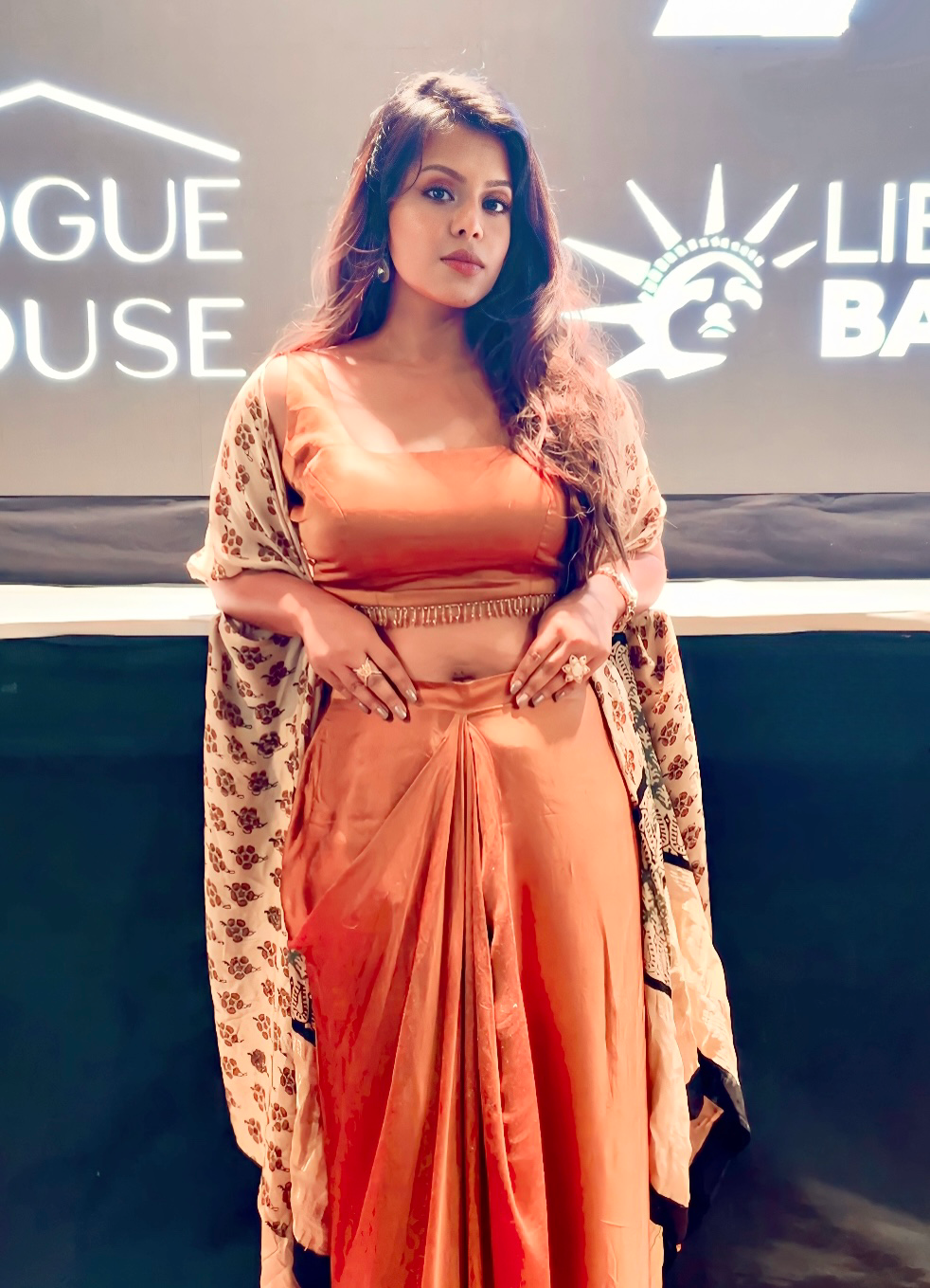 Sharmin Khan stands in front of grey background with orange traditional indian outfit. 