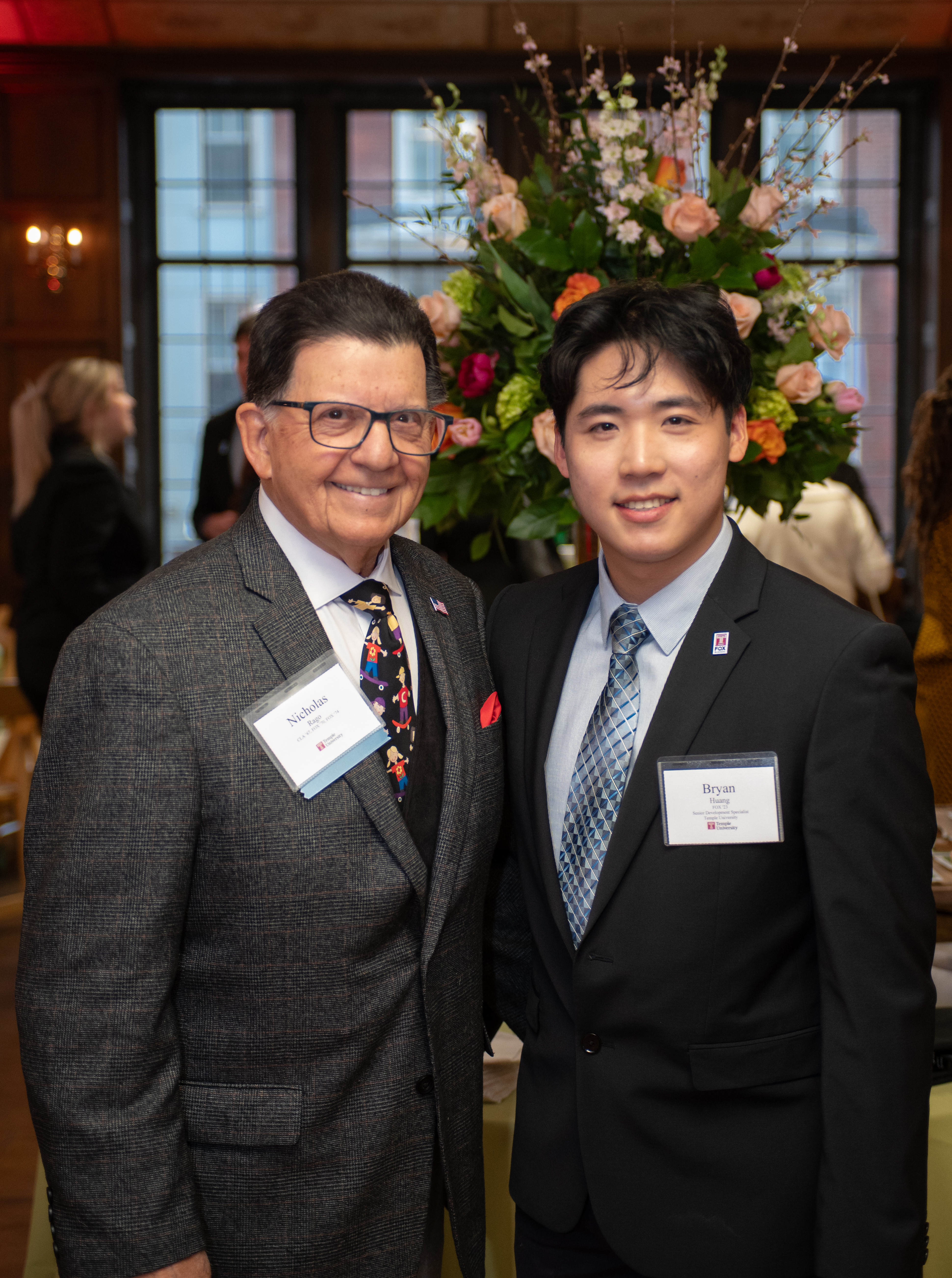 Huang and Rago pictured in suits and name tags standing in front of a floral decoration at the scholarship reception