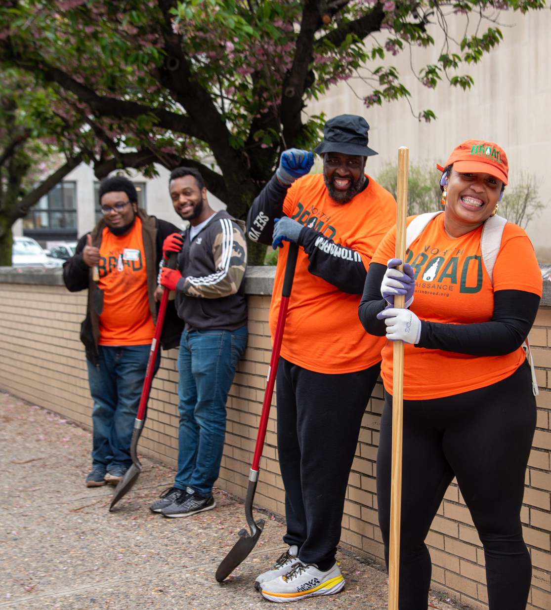 Shalimar and crew clan in orange posing with shovels as they prepare to clean and landscape the area along broad 